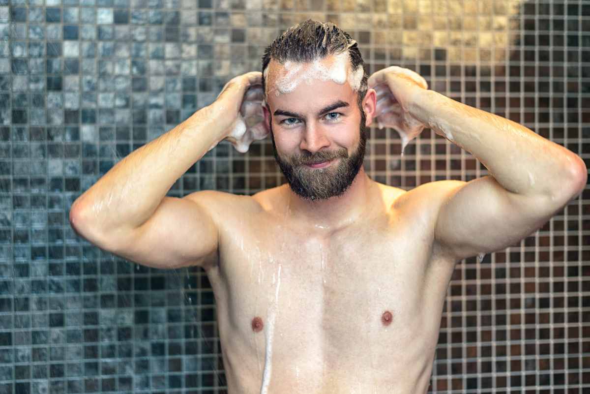Uomo con la barba si lava i capelli con lo shampoo mentre si trova sotto il getto d'acqua sotto la doccia