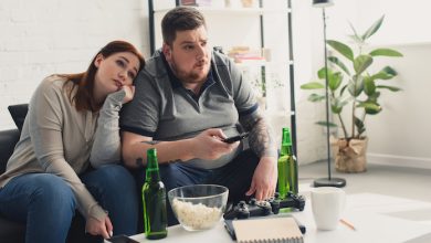 Una donna e un uomo in sovrappeso guardano la tv, bevono alcolici e mangiano popcorn