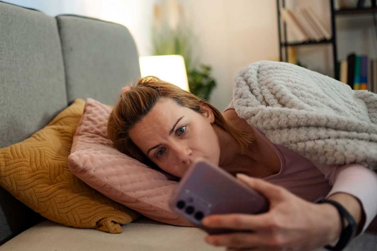 donna che scrolla notizie su smartphone mentre è sdraiata a letto