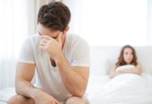 uomo con calo del desiderio sessuale maschile seduto ai piedi del letto mentre si regge la testa e alle spalle una donna sdraiata con le braccia conserte