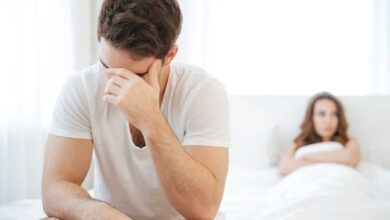 uomo con calo del desiderio sessuale maschile seduto ai piedi del letto mentre si regge la testa e alle spalle una donna sdraiata con le braccia conserte