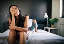 donna seduta sul letto con le mani sulla faccia, sconfortata, mentre il partner è sdraiato alle sue spalle