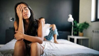 donna seduta sul letto con le mani sulla faccia, sconfortata, mentre il partner è sdraiato alle sue spalle