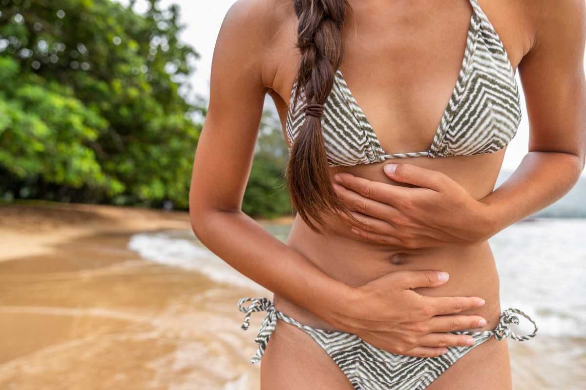 ragazza  sulla spiaggia che si mette le mani sulla pancia perché ha dolore 
