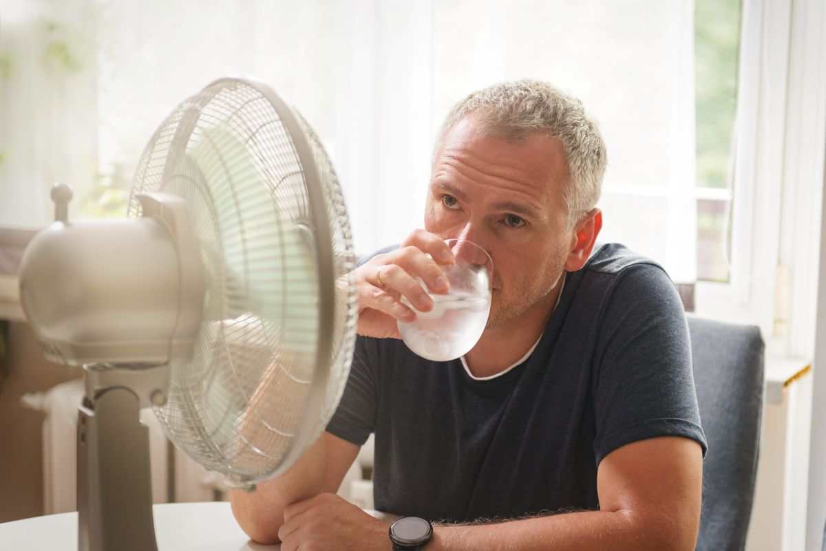 uomo seduto davanti a un ventilatore con un bicchiere di acqua ghiacciata in mano