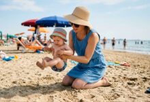 mamma che cerca di appoggiare il bambino piccolo sulla spiggia ma lui ha paura della sabbia e tira su le gambine