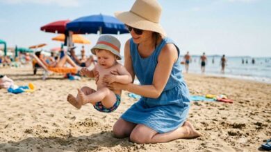 mamma che cerca di appoggiare il bambino piccolo sulla spiggia ma lui ha paura della sabbia e tira su le gambine