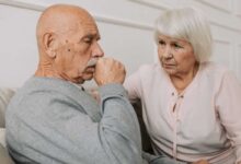 anziano che tossisce portandosi la mano alla bocca con accanto la moglie anziana che lo guarda