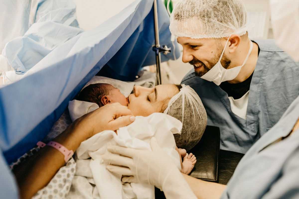 donna che ha appena partorito con taglio cesareo e tiene in braccio il suo bambino con il marito alle spalle che sorride