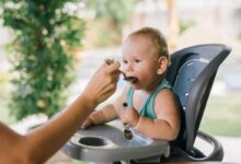 neonato seduto sul seggiolone che ha iniziato lo svezzamento e mangia dal cucchiaino offerto dalla mamma