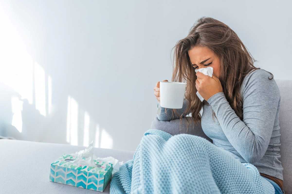 donna con sintomi influenza seduta sul divano mentre si soffia il naso e tiene in mano una tazza