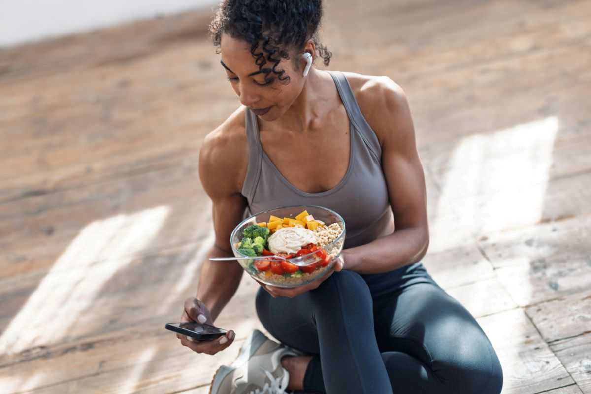 donna sportiva che mangia un piatto sano mentre guarda lo smartphone